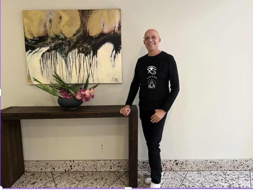 Todd is a fair skin black male, shaved head, clear framed glasses, bright blue eyes is modeling the look within long sleeve black shirt with an icon of they all seeing eye of horus over text look within over an image of a person meditating with rays of aura around the their body.