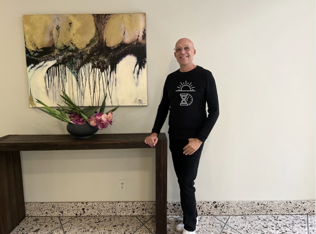 Todd is a fair skin black male, shaved head, clear framed glasses, bright blue eyes is modeling the Sun will come out tomorrow black long sleeve shirt.  In white an image of a rising sun over and hourglass with a partial view of a clock at 5 am.