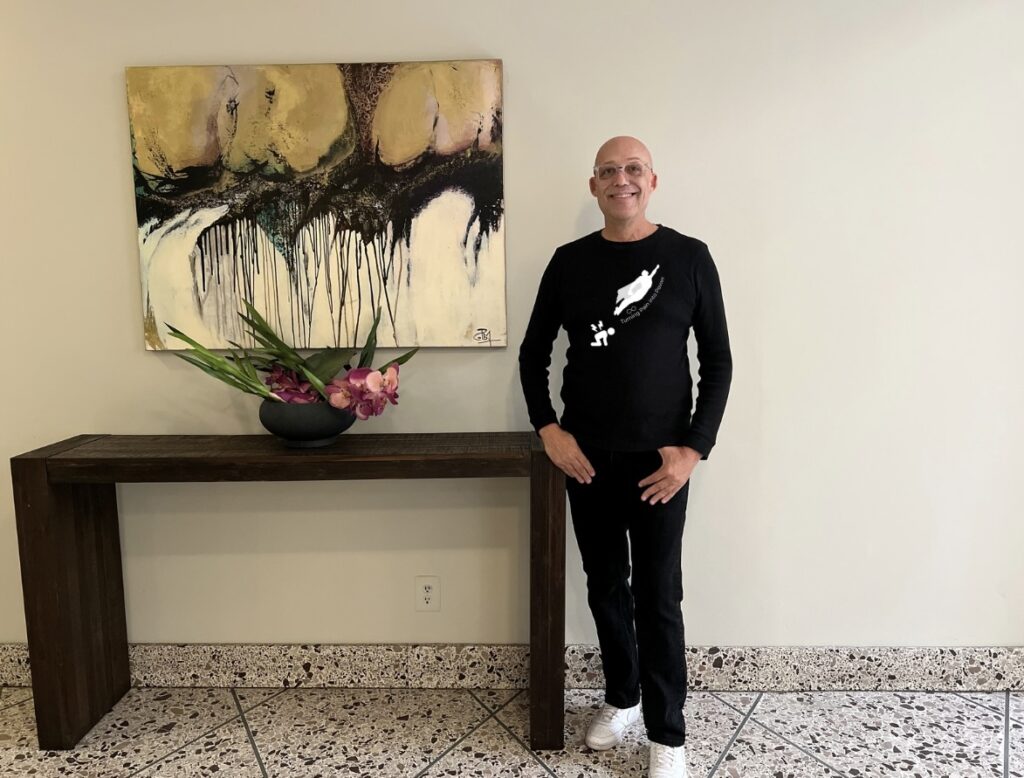 Todd is a fair skin black male, shaved head, clear framed glasses, bright blue eyes is modeling the look within long sleeve black shirt with an icons of a man on his hands and knees with pulsing back pain, just above diagonally a man in a superman cape simulating flying, text: Turning pain into power.  